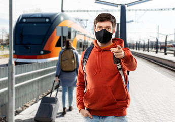 travel, tourism and pandemic concept - young man with backpack in face protective medical mask pointing finger to camera over train on railway station in city of tallinn, estonia on background