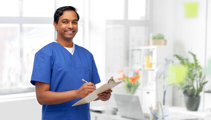 healthcare, profession and medicine concept - happy smiling indian doctor or male nurse in blue uniform writing report on clipboard over medical office at hospital background