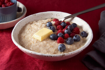 Oatmeal porridge with blueberry, bilberry. Breakfast in white bowl and spoon with fresh berries and butter on dark red cranberry linen tablecloth. Bright color of textile. Side view, copy space
