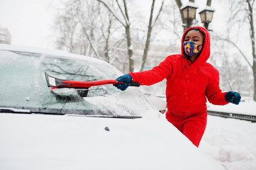Fototapeta na wymiar African american woman in red hoodie and face mask clean car from snow in winter day.
