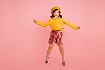 Full length shot of cute female kid in yellow beret. Studio photo of excited preteen girl jumping on pink background.