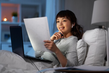 technology, remote job and people concept - happy smiling young asian woman with laptop computer, papers and earphones working in bed at home at night