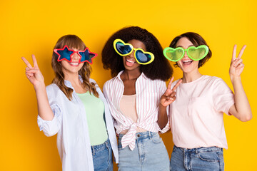 Portrait of attractive trendy cheerful girls embracing showing v-sign wearing large specs isolated over bright yellow color background
