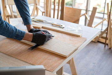 Professional carpenter man wearing glove using pencil and ruler to draw the line on the wood piece in the modern wood workplace. There is goggles and saw on the table. Furniture DIY concept