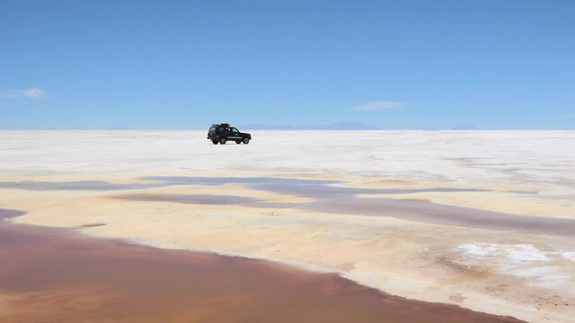 Black jeep drives over flat white crust of salt with a body of brown water at the foreground. Uyuni Salt Flats, Bolivian highlands.