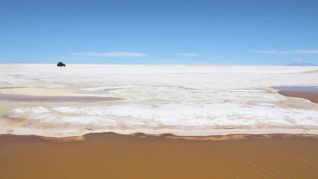 Little black car far away in a white desert of Salt Flats of Salar de Uyuni. Brown water basin at the foreground.
