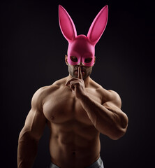 Portrait of shirtless handsome muscular man, athlete with perfect built body wearing pink rabbit, bunny mask stands with naked chest and gesturing silence, secret sign with finger on dark background - 417818217