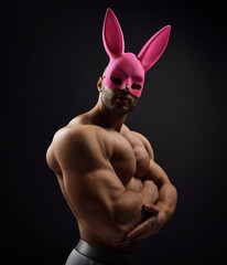 Portrait of shirtless handsome muscular man, athlete with perfect built body wearing pink rabbit mask standing naked sideways to camera demonstrating his biceps and chest over dark background - 417818445
