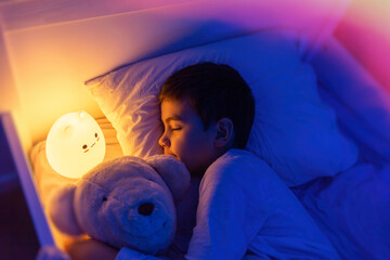Little preschool kid boy sleeping in bed with colorful night led lamp. School child dreaming and holding plush toy. Kid angry of darkness.