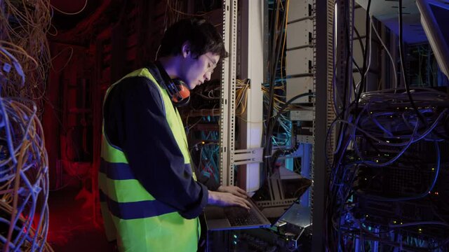 Medium shot of young Asian IT engineer wearing special uniform and glasses standing in data center and talking to colleague by cellphone