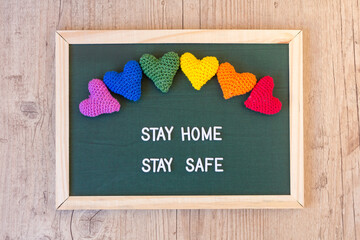 Blackboard with decorative hearts in the colors of the rainbow with the positive message: stay home, stay safe. Wooden background.