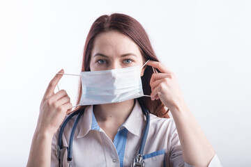 Portrait of a female doctor in a white uniform who puts on a medical mask in the studio on a white background