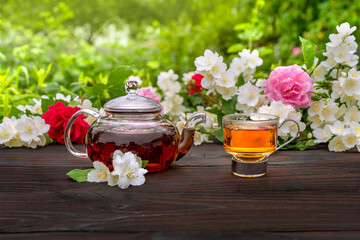 A steamed teapot and an elegant glass cup among flowering rose and jasmine bushes. Outdoor, picnic, brunch. Green leaves background in blur.