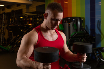 A strong male athlete in a red T-shirt tensely holds dumbbells and looks down standing in the gym. High quality photo