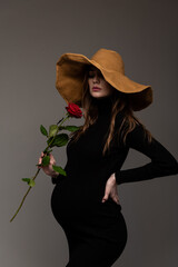 Pregnant girl in black tight dress. In a big hat. With a rose in my hand. Hand on the waist. Looking down. Grey background.