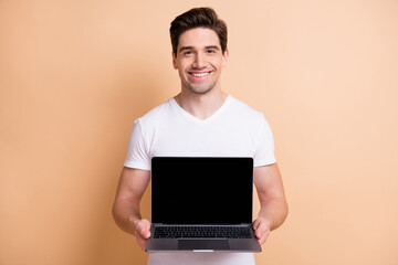 Portrait of nice young man toothy smile arms hold netbook showing display isolated on beige color background