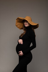 Pregnant girl in black tight dress. In a big hat. Hands on belly. Grey background.