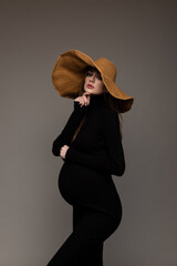 Pregnant girl in black tight dress. In a big hat. Hand to face. Grey background.