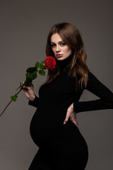 Pregnant girl in black tight dress. With a rose in my hand. Hand on the waist.  Grey background.