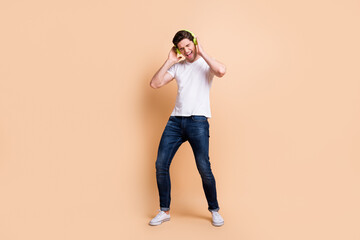 Fototapeta na wymiar Full size photo of optimistic brunet man listen music wear white t-shirt jeans sneakers isolated on beige color background