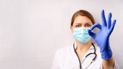 Female doctor in mask and blue medical gloves shows ok with fingers