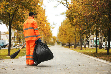 Street cleaner with broom and garbage bag outdoors on autumn day, back view