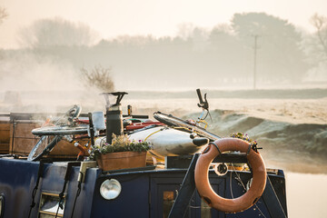 Smoke coming from chimney of wood log burning narrow canal boat on misty morning river scene with lifebuoy orange preserver ring canoe and mountain bike on roof.