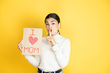 Beautiful woman celebrating mothers day holding poster love mom message asking to be quiet with finger on lips. Silence and secret concept.