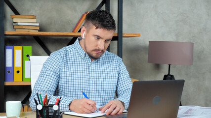 Bearded focused businessman at home office wear wireless earphones looks laptop computer screen display writes information notebook, remote online distance video call conference webcam virtual meeting