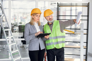 Portrait of pretty confident mature blond business woman inspecting with elderly bearded engineer or contractor building works on construction site indoors