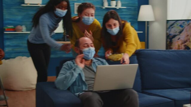 Cheerful happy multi ethnic friends looking at photo on laptop having fun together keeping social distance wearing face mask preventing coronavirus spreading. Diverse people enjoying new normal party