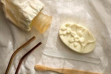 Sustainable toiletries and utensils on linen cloth: metal straws, wooden toothbrush, glass nail file, bar of soap and menstrual cup. Top view.