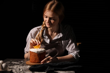 Young woman decorating traditional Easter cake at table against black background. Space for text