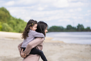 Fototapeta premium Happy woman and little girl playing together outside on scenic spring sandy beach. Happy white family of young mother and daughter enjoying warm spring nature