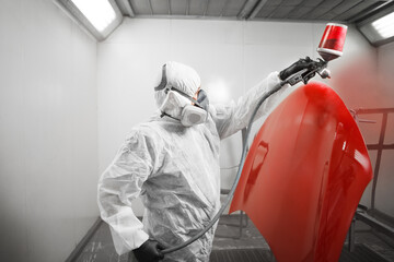 Automobile repairman painter in mask and protective workwear painting car body in paint chamber.