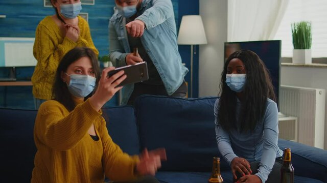 Happy friends from diverse cultures and races taking selfie wearing protection masks during new normal party having fun together keeping social distance, spending free time in living room