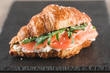 One croissant sandwich with salmon, ricotta cheese and arugula on a black slate board on a dark background.