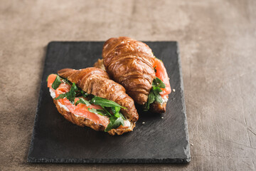 Two croissant sandwich with salmon, ricotta cheese and arugula on a black slate board on a dark background.