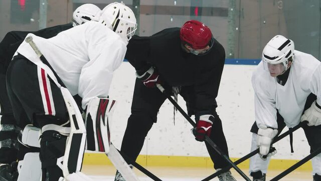 Tilting-up shot of professional hockey coach in red helmet and young male players standing in circle on ice while training, discussing hockey tricks and techniques