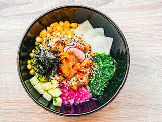 Poke bowl with salmon, avocado, radish, corn, seaweed, peas, nori, daikon, sesame seeds. Asian food in a black plate on a wooden table in a cafe, restaurant. Top view