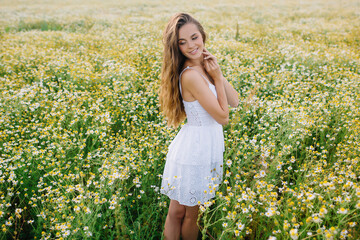 Naklejka na ściany i meble Very beautiful woman model stands in a field with daisies in a white dress with long hair