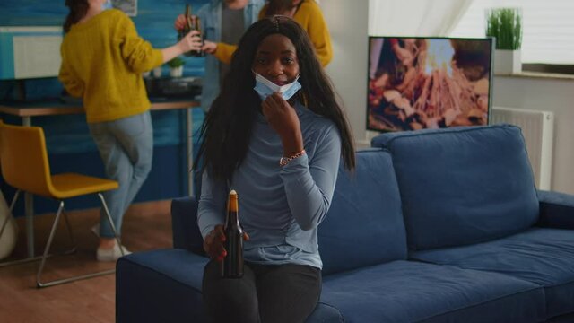 African woman keeping social distancing wearing face mask while meeting with friends to prevent spread of coronavirus holding beer bottle looking at camera sitting on couch, covid 19 outbreak