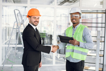 Smiling multiracial men in protective helmets and glasses greeting each other with hand shaking. Two business partners working on common construction project.