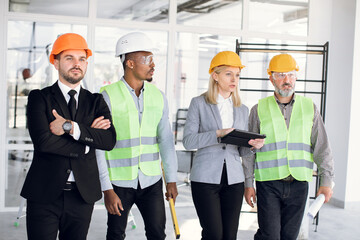 Group of multiethnic confident male and female architects and engineers in hardhats inspecting unfinished office building together and discussing plan details