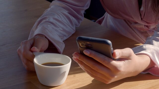 Young mixed-race female wearing velvet pink robe. A cup of hot freshly brewed coffee is on the wooden table. The woman is interacting with her phone. Sunny day. Close up no face high-resolution image