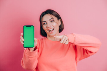 Naklejka premium Look at this cell phone! Pleased happy woman points with index finger at blank screen, shows modern device, happy surprised emotions.