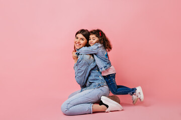 Obraz na płótnie Canvas Pleasant kid embracing mother on pink background. Studio shot of blissful mom and little daughter in jeans.