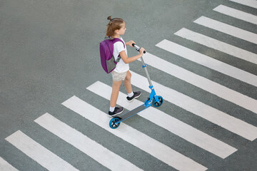 Schoolgirl carrying scooter and crossing road on way to school. Zebra traffic walk way in the city. Pedestrian passing a crosswalk. Stylish child walking with backpack. Safety concept for road users © Igor
