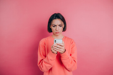 Young woman is feeling sad unhappy angry while reading sms using her mobile phone, over pink background