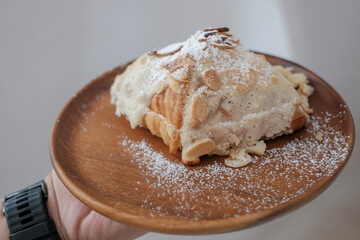  Fresh croissant breakfast. Croissant with almond, classic traditional French breakfast on white plate, top of icing sugar and almond slide. Light brown wood background.